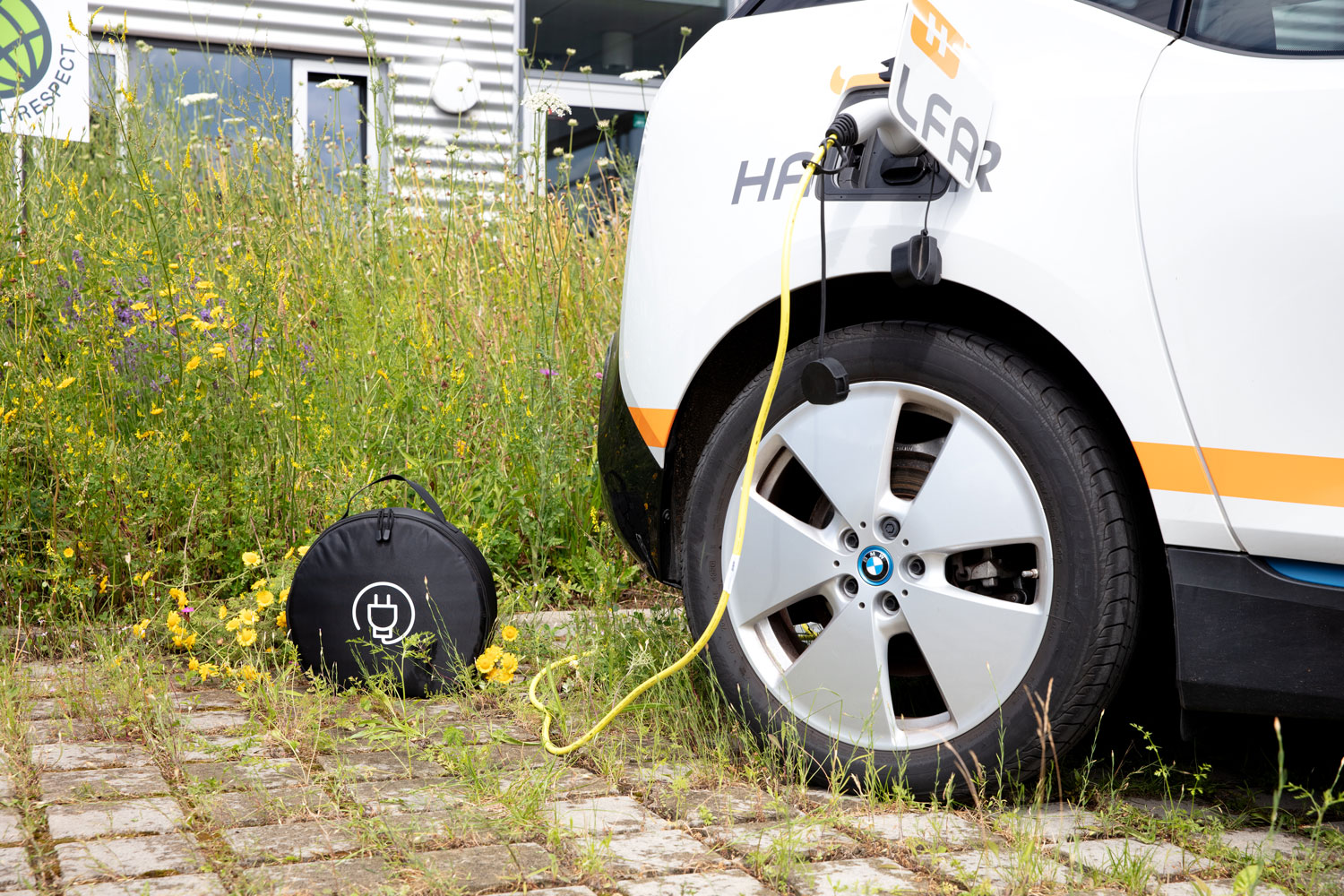 Halfar E-Auto mit Ladekabel vor einer Grünfläche und dem Firmengebäude